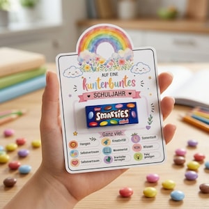 Puede incluir: Una tarjeta rectangular blanca con una ilustración colorida de un arcoíris y nubes en la parte superior, con el texto "AUF EINE kunterbuntes SCHULJAHR" y una caja pequeña de caramelos Smarties azul en el centro, rodeada de etiquetas en alemán.