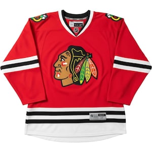May include: A red hockey jersey featuring the Chicago Blackhawks logo on the chest, with black and white stripes on the sleeves and hem. The V-neck collar is white and black, and the shoulders display team logos. The jersey includes an NHL logo patch.