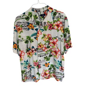 May include: White short-sleeved shirt with a tropical floral print. The shirt features a vibrant pattern of red, yellow, and pink hibiscus flowers, palm trees, and island scenery. The shirt is hanging on a metal hanger.
