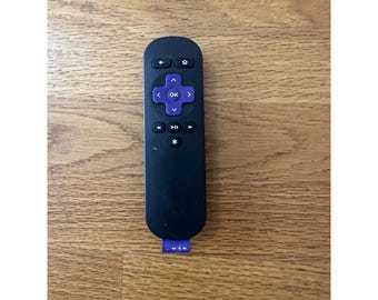 May include: A black Roku remote control with purple directional buttons and a purple tab at the bottom. The remote has buttons for power, home, back, and play/pause. The remote is lying on a wooden surface.