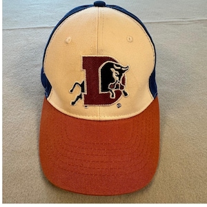 May include: A baseball cap with a curved brim. The cap features a cream-colored crown, a burnt orange brim, and navy blue side panels. A maroon letter "D" with a bull logo is embroidered on the front.