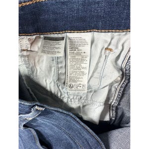 May include: Close-up of the inside of a pair of blue denim jeans. The jeans' interior features a white tag with text in multiple languages, including English, Spanish, and Japanese. The jeans are folded, showing the stitching and fabric details.