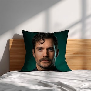 May include: A decorative square pillow featuring a portrait of a man with dark hair, a mustache, and a beard. The pillow has a dark green background and is placed on a bed with white sheets.