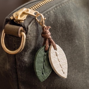 May include: An olive green waxed canvas bag featuring a gold-toned metal zipper closure and a D-ring. Attached to the zipper pull is a decorative charm consisting of two small, leaf-shaped leather pieces in dark green and cream with stitching.