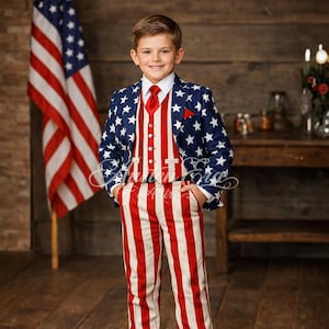 Op de afbeelding: Een kind draagt een patriottisch pak met een marineblauwe jas bedekt met witte sterren. Het vest is rood en wit gestreept, passend bij de broek. Een rode stropdas en pochet maken de outfit compleet, met een Amerikaanse vlag op de achtergrond.