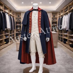 Puede incluir: Un disfraz histórico en un maniquí. El atuendo incluye una capa azul marino con forro rojo, un chaleco color crema con botones dorados y calzones a juego. El disfraz se exhibe en una habitación bien iluminada con percheros.