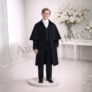 Op de afbeelding: Een jong persoon draagt een zwart formeel pak met een bijpassende cape. Het pak bestaat uit een vest, een pantalon en een lange jas. Een wit overhemd en zwarte schoenen maken de outfit compleet. De achtergrond is een heldere witte kamer met een groot bloemstuk.