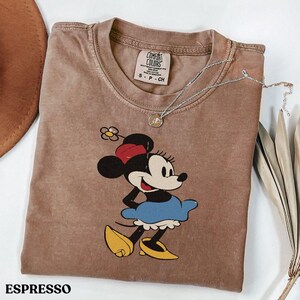Puede incluir: Camiseta color espresso con un gr&aacute;fico de Minnie Mouse. La camiseta tiene cuello redondo y mangas cortas. Un collar plateado con un colgante est&aacute; drapeado sobre la camiseta. La camiseta est&aacute; doblada sobre una superficie blanca. La palabra "ESPRESSO" est&aacute; impresa en la parte inferior.