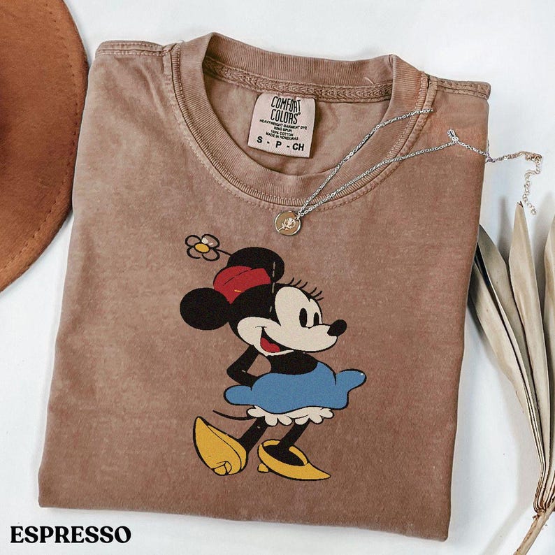 Puede incluir: Camiseta color espresso con un gr&aacute;fico de Minnie Mouse. La camiseta tiene cuello redondo y mangas cortas. Un collar plateado con un colgante est&aacute; drapeado sobre la camiseta. La camiseta est&aacute; doblada sobre una superficie blanca. La palabra "ESPRESSO" est&aacute; impresa en la parte inferior.