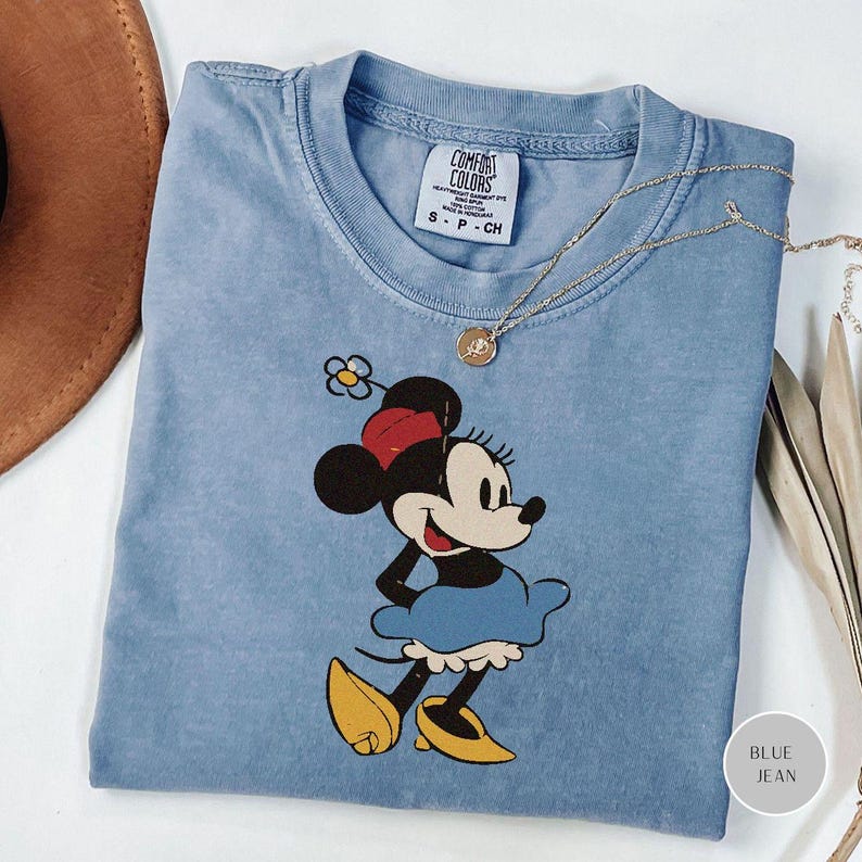Puede incluir: Camiseta Comfort Colors azul claro con un gr&aacute;fico de Minnie Mouse con un vestido azul, zapatos amarillos y un lazo rojo. Un collar dorado con un colgante est&aacute; drapeado sobre la camisa. La camisa tiene el texto "S - P - CH".