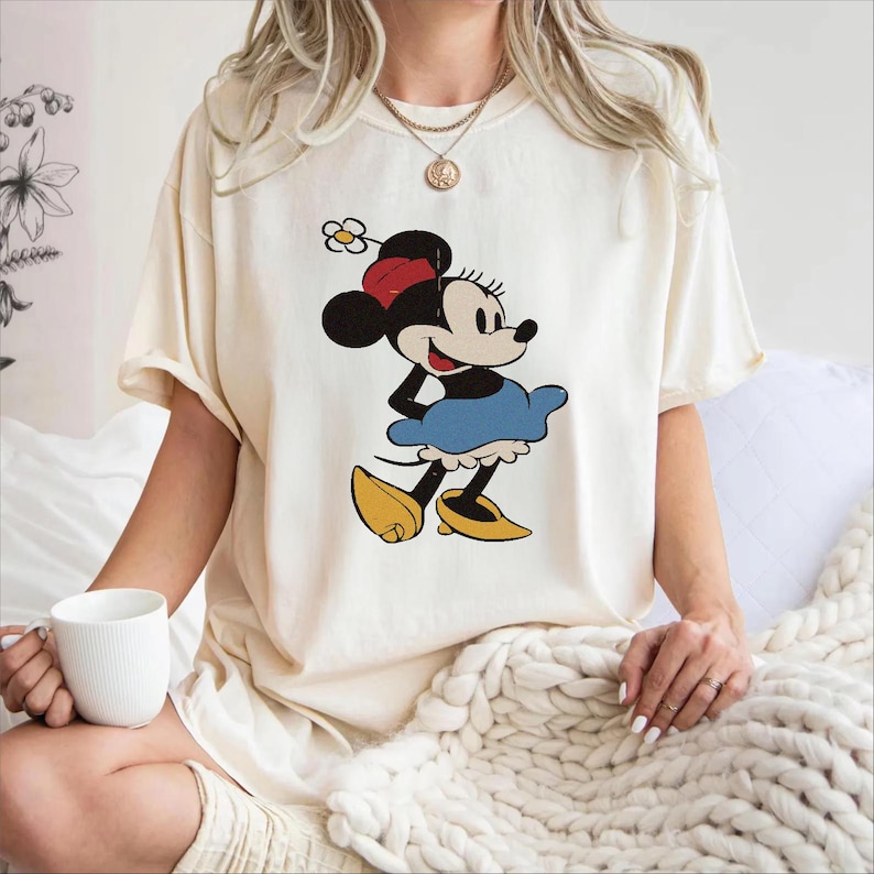 Puede incluir: Camiseta color crema con una imagen de dibujos animados cl&aacute;sica de Minnie Mouse. El gr&aacute;fico muestra a Minnie con una falda azul, zapatos amarillos y un sombrero rojo y negro. La camiseta tiene mangas cortas y un corte holgado.