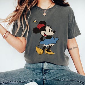 Puede incluir: Camiseta gris oscuro con un dise&ntilde;o vintage de Minnie Mouse. Minnie aparece con su atuendo cl&aacute;sico: lazo rojo, falda azul y zapatos amarillos. La camiseta tiene un aspecto ligeramente descolorido, lo que sugiere un estilo vintage.