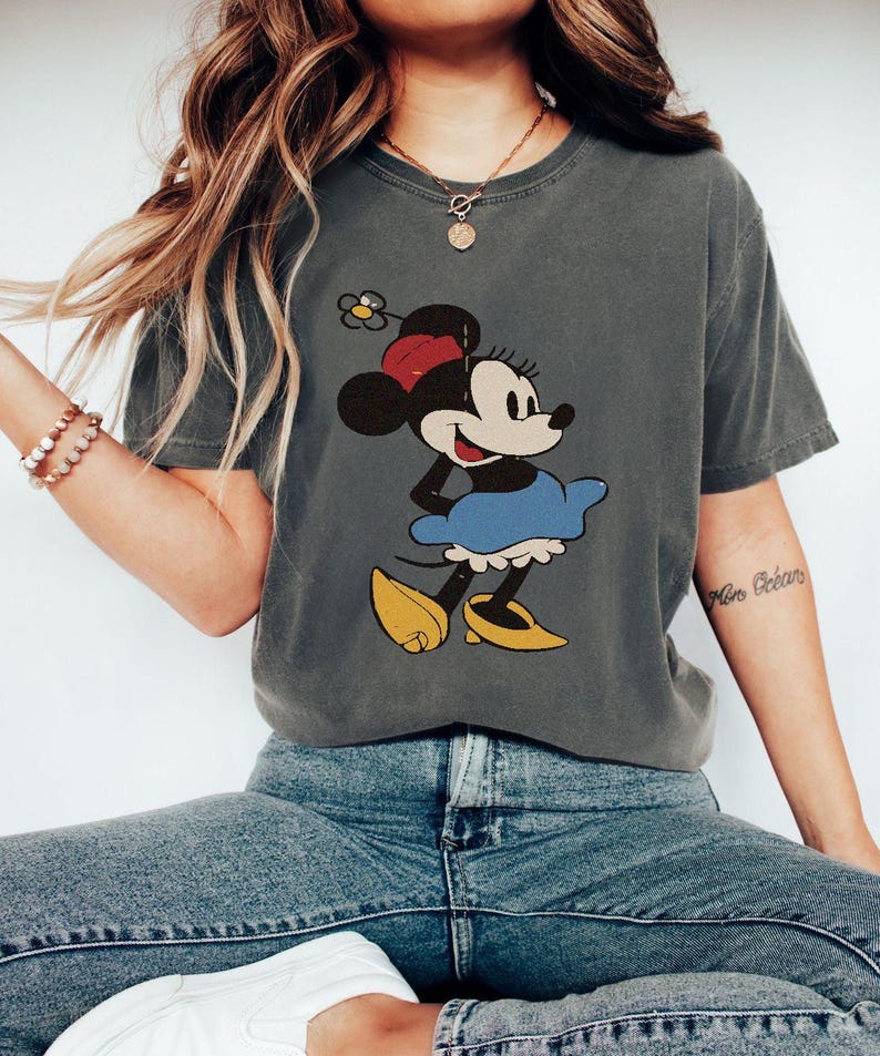 Puede incluir: Camiseta gris oscuro con un dise&ntilde;o vintage de Minnie Mouse. Minnie aparece con su atuendo cl&aacute;sico: lazo rojo, falda azul y zapatos amarillos. La camiseta tiene un aspecto ligeramente descolorido, lo que sugiere un estilo vintage.
