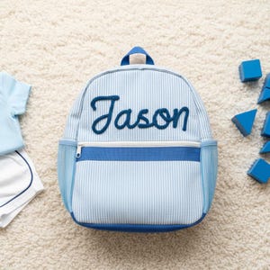 May include: Light blue and white striped backpack with the name "Jason" embroidered in dark blue script. The backpack has a front zipper pocket, mesh side pockets, and a top handle. Blue toy blocks and a blue shirt and shorts set are in the background.