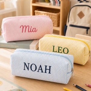 May include: Three rectangular pencil cases with a seersucker striped pattern. The top case is pink with the name Mia embroidered in red. The middle case is yellow with the name LEO in green. The bottom case is blue with the name NOAH in navy.