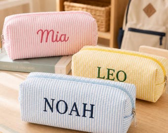 May include: Three rectangular pencil cases with a seersucker striped pattern. The top case is pink with the name Mia embroidered in red. The middle case is yellow with the name LEO in green. The bottom case is blue with the name NOAH in navy.