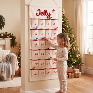 Könnte beinhalten: Ein weißer Wand-Adventskalender aus Stoff mit vierundzwanzig Taschen, die mit roten Zahlen von 1 bis 24 versehen sind. Oben steht der Name Jelly in roter Schreibschrift neben einem kleinen Schneemann-Symbol. Jede Tasche hat einen roten Rand.