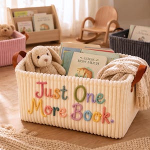 May include: A cream-colored corduroy storage basket with brown leather handles. The front features the embroidered text "Just One More Book." in colorful yarn letters. Inside the basket sits a plush tan bunny toy, several children's books, and a folded cream-colored knit blanket with fringe edges.