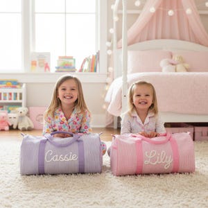 Puede incluir: Dos bolsas de lona, una lavanda y otra rosa, están sobre una alfombra blanca. La bolsa lavanda tiene bordado el nombre "Cassie", y la rosa "Lily". Ambas bolsas tienen rayas verticales y asas a juego.