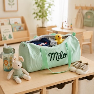 May include: A light green duffel bag with white pinstripes and the name Milo embroidered in dark green script. The bag is open, revealing a blue shirt and a small yellow duck toy. A stuffed rabbit, a water bottle, and two small beige trainers sit nearby.