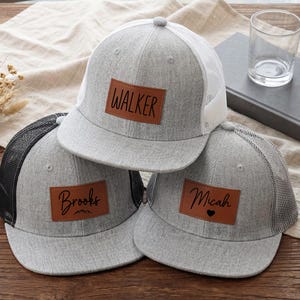 May include: Three gray trucker hats with personalized leather patches. One hat has "WALKER" on the patch, another has "Brooks", and the third has "Micah" with a heart symbol. The hats have mesh backs and are displayed on a wooden surface.