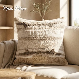 May include: A square decorative pillow with a textured, striped design in shades of cream and brown. The pillow is placed on a beige sofa. The text "Mimi Pillow" is in the upper left corner. The pillow has a soft, plush appearance.