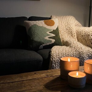 May include: A cozy living room scene featuring a dark gray sofa, a decorative pillow with a mountain and sun design, and a cream-colored chunky knit blanket. Three lit candles in ceramic holders sit on a wooden table.