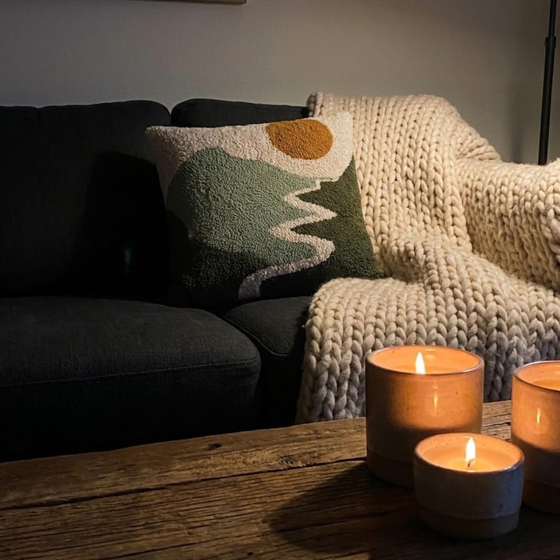 May include: A cozy living room scene featuring a dark gray sofa, a decorative pillow with a mountain and sun design, and a cream-colored chunky knit blanket. Three lit candles in ceramic holders sit on a wooden table.