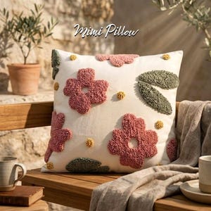 Könnte beinhalten: Cremefarbenes Mimi-Kissen mit gestickten rosa Blumen, grünen Blättern und gelben Akzenten. Das Kissen liegt auf einer Holzbank mit einer neutralfarbenen Decke und einer Tasse. Der Text "Mimi Pillow" steht oben.