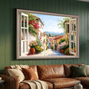 Könnte beinhalten: Ein gerahmtes Kunstwerk, das eine lebendige italienische Straßenszene durch ein offenes Fenster zeigt. Das Kunstwerk zeigt eine Kopfsteinpflasterstraße mit Gebäuden, Blumen und Terrakottatöpfen. Der Fensterrahmen ist weiß, vor einer grünen Wand.