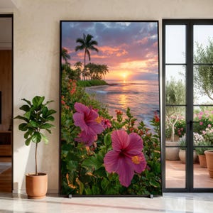 May include: A framed print depicting a tropical beach at sunset. The image shows bright pink hibiscus flowers, green foliage, palm trees, and the ocean under a colourful sky. The print is displayed indoors, next to a potted plant.