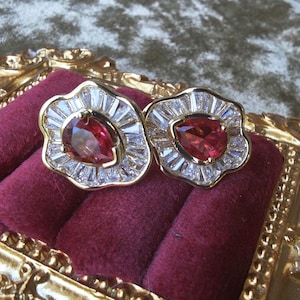 Op de afbeelding: Een paar goudkleurige oorbellen met een druppelvormige rode edelsteen in het midden, omringd door een halo van witte baguette-geslepen stenen. De oorbellen liggen op een dieprode fluwelen ondergrond.