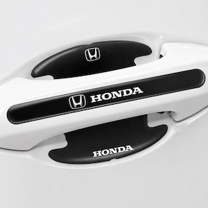 以下が含まれることがあります： 白の車のドアハンドルのクローズアップ。黒いアクセントが付いています。ハンドルにはホンダのロゴと「HONDA」の文字が白で表示されています。デザインには、ドアハンドルのグリップ用の黒いインサートが含まれています。