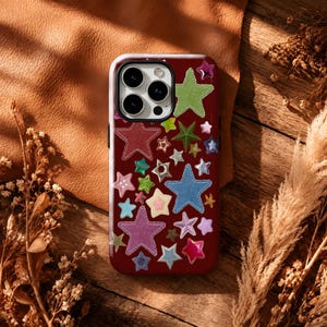 May include: A dark red smartphone case featuring a scattered pattern of various star shapes in multiple colors, including blue, green, pink, and silver. The stars have different textures and finishes, some appearing as fabric patches with stitching.