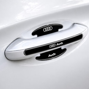 以下が含まれることがあります： 黒いアクセントが付いた白い車のドアハンドルのクローズアップ。ハンドルにはアウディのロゴと「Audi」の文字が白で表示されています。ハンドルは白い車のドアにあります。