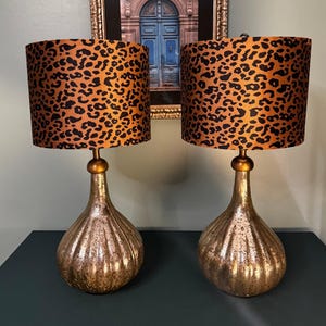 May include: A pair of table lamps featuring cylindrical shades with a black and orange leopard print pattern. The bases are made of metallic, mercury-glass style material with a ribbed, teardrop shape and a smooth, rounded gold-toned neck.