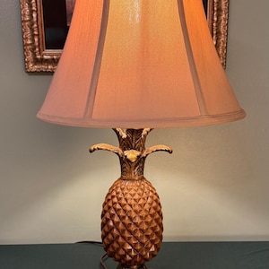 May include: A table lamp featuring a base shaped like a textured, golden-brown pineapple with a square pedestal. The lamp is topped with a conical, light-brown fabric lampshade that emits a warm, soft glow, casting light downward over the pineapple base.
