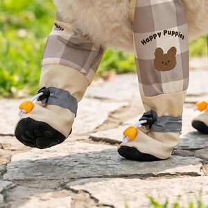 May include: Tan and brown plaid dog pants with matching booties. The pants have the words "Happy Puppies" and a bear graphic. The booties have black soles, reflective gray bands, and orange toggles.