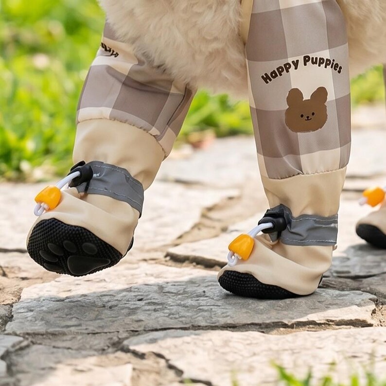 May include: Tan and brown plaid dog pants with matching booties. The pants have the words "Happy Puppies" and a bear graphic. The booties have black soles, reflective gray bands, and orange toggles.