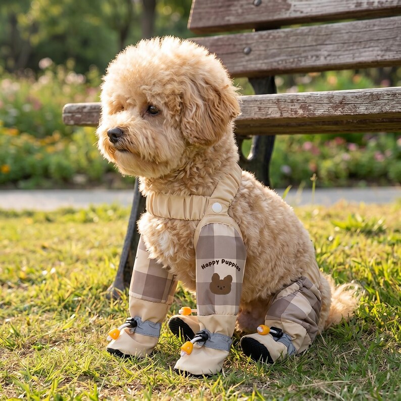 May include: A small, light brown dog wearing a beige and brown plaid jumpsuit with matching booties. The jumpsuit has the words "Happy Puppies" and a bear graphic on the leg. The dog is sitting on green grass near a wooden bench.