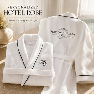 May include: Two white waffle-knit hotel robes with black piping trim. One robe is folded, featuring the embroidered monogram AE on the chest. The other robe is displayed upright, showing the text MAISON AURELIA Spa printed on the back.