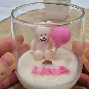 Peut inclure: Un bougeoir en verre transparent avec un ours en peluche rose, un ballon rose et le nom "LINA" en lettres roses. La bougie est blanche et surmontée d'un petit morceau de chocolat.