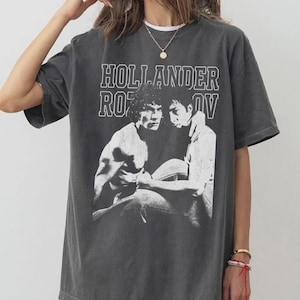 May include: Dark gray t-shirt featuring a black and white graphic of two men and the text "HOLLANDER ROV". The shirt has a vintage, worn look and a crew neck.