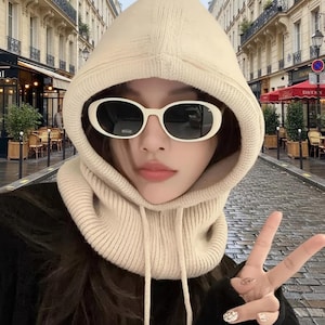 May include: A beige knit hood with a built-in scarf, worn with white oval sunglasses. The hood is made of a soft, textured material and provides warmth and style. The sunglasses have dark lenses.