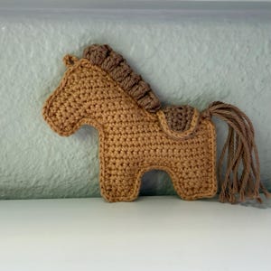 May include: A crocheted horse figurine in shades of brown. The horse has a detailed mane, tail, and saddle. The handmade craft is displayed against a neutral background, showcasing the texture and craftsmanship of the yarn.