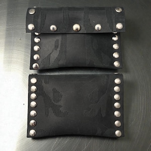 以下が含まれることがあります： シルバー色のスタッドが付いた2つの黒い財布。上部の財布はフラップ開閉式で、わずかに開いています。財布には微妙な模様があり、テクスチャード加工された素材で作られています。財布は金属の表面に表示されています。