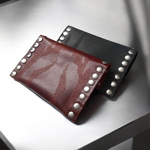 May include: Two leather wrist cuffs, one burgundy and one black, are displayed. Each cuff is adorned with a row of silver-colored studs along the top edge. The burgundy cuff has a textured surface, while the black cuff appears smooth.