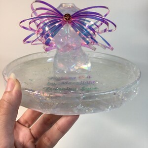 May include: A clear iridescent resin trinket dish with a glittery finish. The dish features a small iridescent resin figurine with a pink and purple iridescent bow. The dish has the text "Catching Drift by Moonlight, Turning Love" written on the bottom.