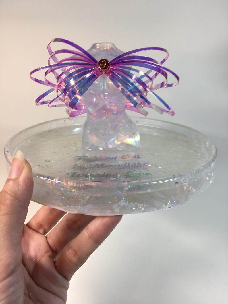 May include: A clear iridescent resin trinket dish with a glittery finish. The dish features a small iridescent resin figurine with a pink and purple iridescent bow. The dish has the text "Catching Drift by Moonlight, Turning Love" written on the bottom.
