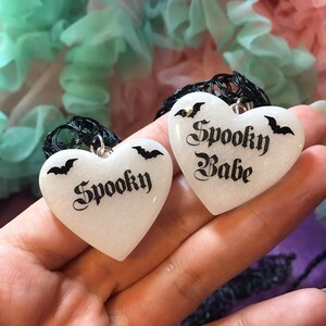 May include: Two white heart-shaped pendants with black bats and the words "Spooky" and "Spooky Babe" in black script.